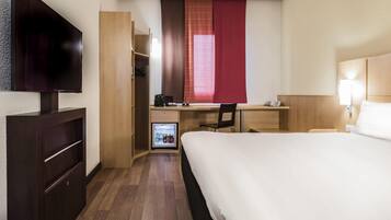 Una habitación de hotel con cama, escritorio y silla, televisor, armario y minibar.