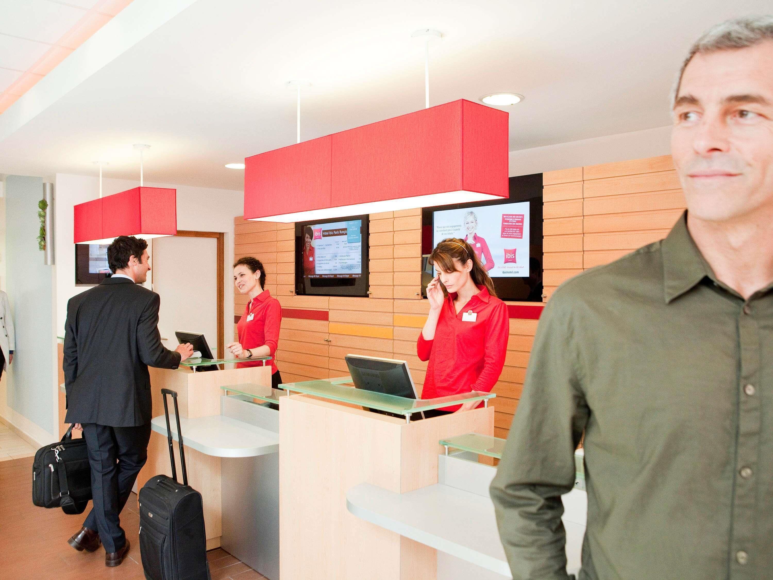 A hotel reception area with a modern design, featuring a reception desk, a digital screen displaying information, and a wooden panel wall.