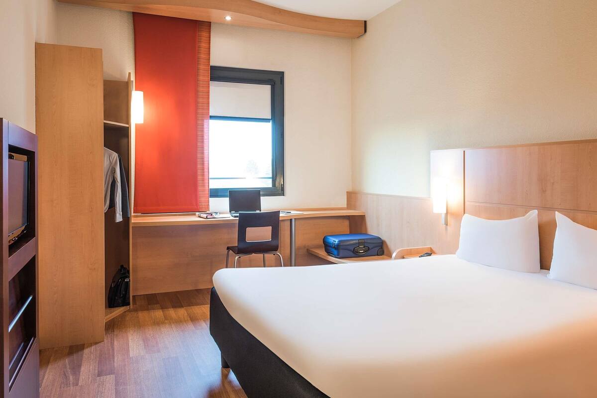 Hotelkamer met een bed, een bureau met laptop, een stoel, een kledingkast en een raam met gordijnen.