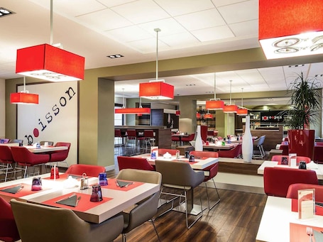 Un interno ristorante moderno con schema di colori rosso e bianco, diversi tavoli e lampade a sospensione rosse.. ibis Milano Malpensa Aeroporto