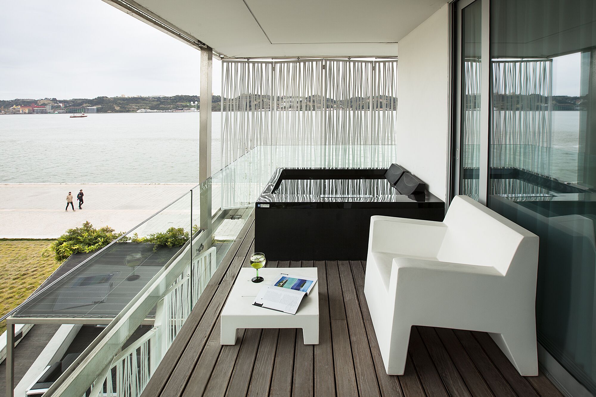 Un balcón con un sofá blanco, una mesita con una revista y vistas al mar y edificios lejanos.