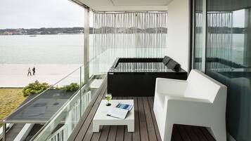 Un balcón con un sofá blanco, una mesita con una revista y vistas al mar y edificios lejanos.
