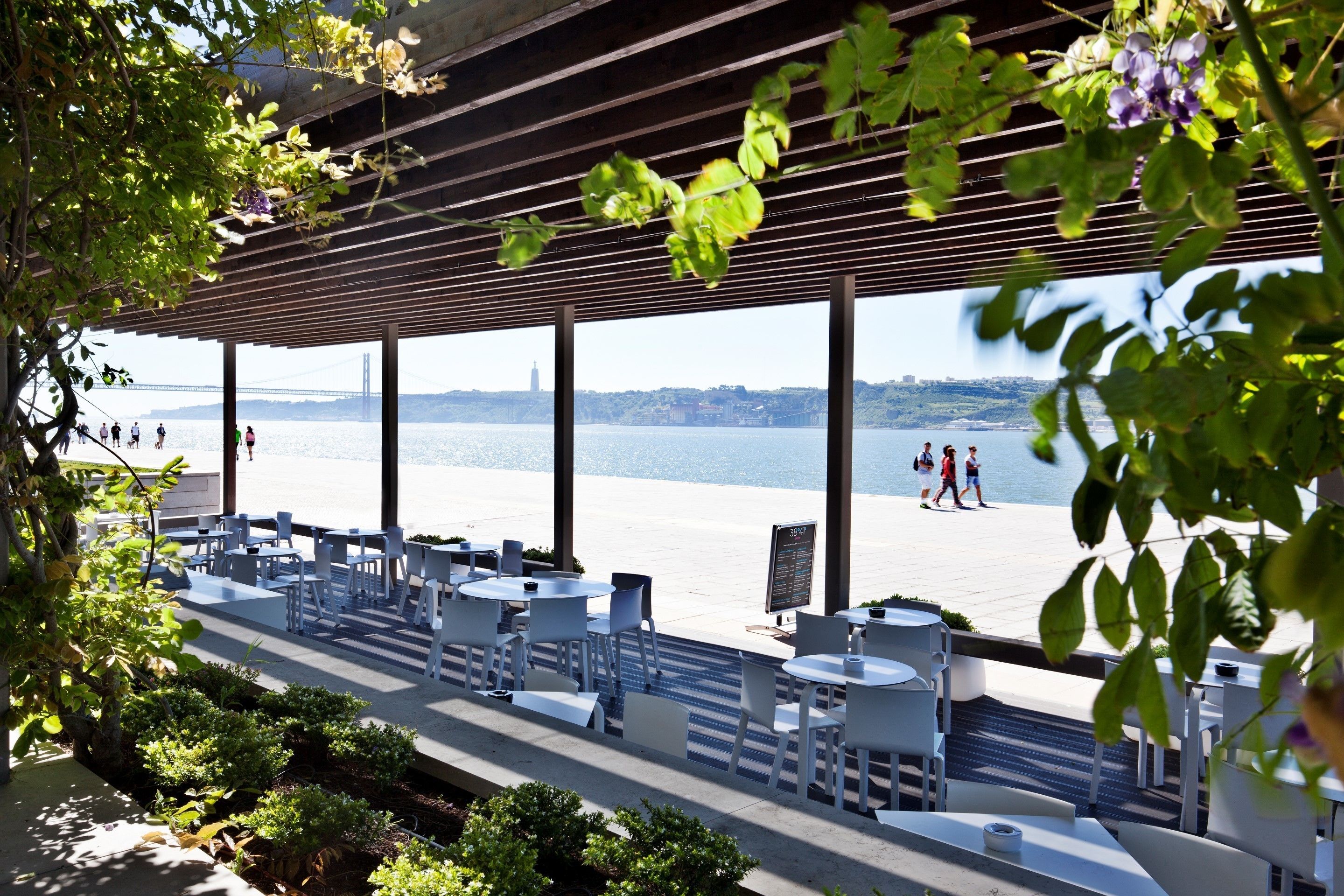 Zona de estar al aire libre con mesas y sillas, con vista a un muelle donde pasean personas.