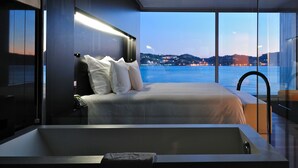 Un dormitorio moderno con una cama grande, un baño con paredes de vidrio y vistas a un cuerpo de agua al atardecer.