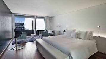 A modern hotel room with a large bed, a desk, and a chair. There is a balcony with a view of the sea.