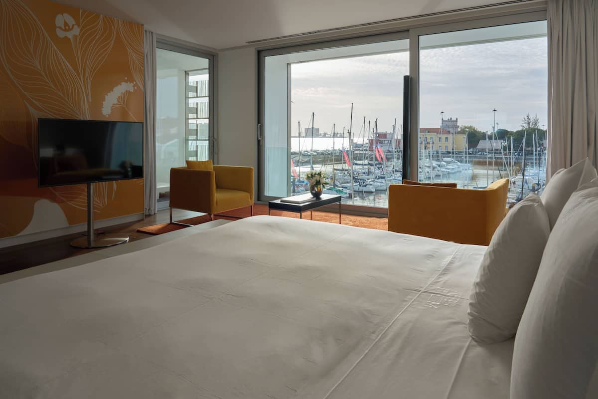 Una habitación de hotel con una cama grande, un televisor, dos sillones amarillos, una mesita con un florero y vista a un puerto deportivo con barcos.