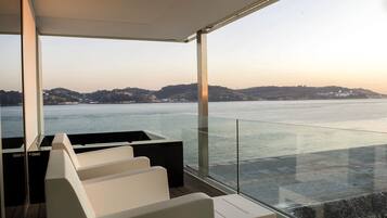 A modern outdoor seating area with a glass balustrade overlooking a body of water and distant hills.