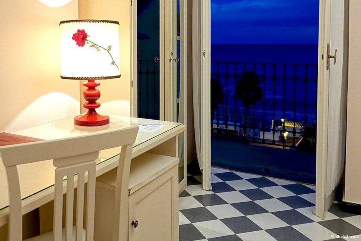 A hotel room with a desk, a lamp with a flower design, a laptop, and a view of a balcony with a checkered floor and a railing.