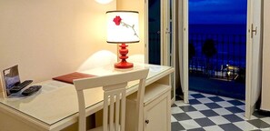A hotel room with a desk, a lamp with a flower design, a laptop, and a view of a balcony with a checkered floor and a railing. - Hotel SplendidMare (Laigueglia)