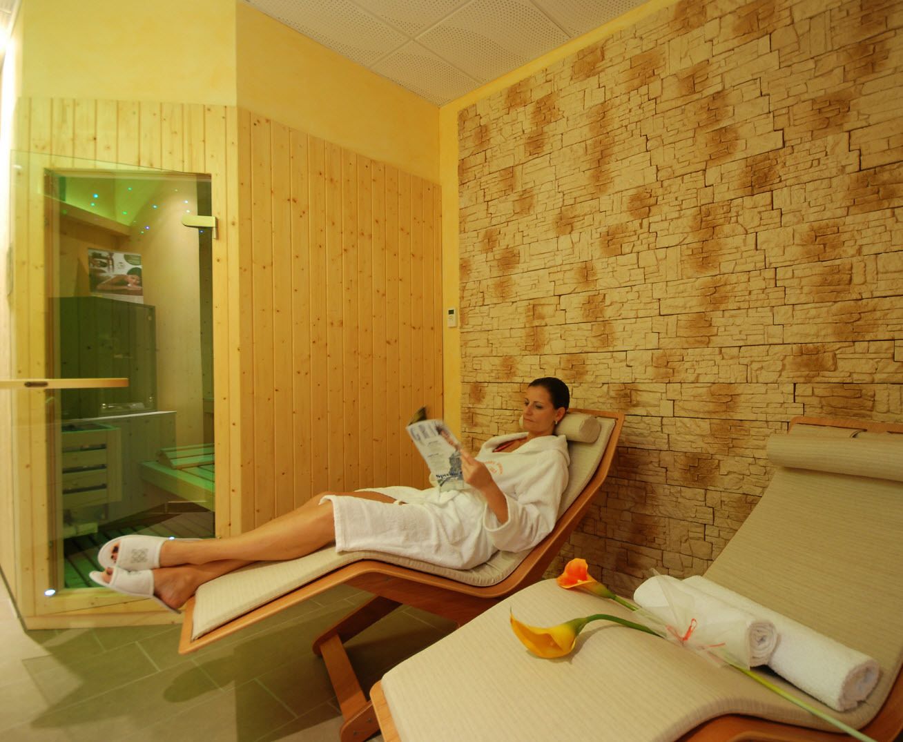 Un cuarto de spa con paneles de madera y una pared con textura de piedra. Una persona está recostada en una camilla tomando un libro.
