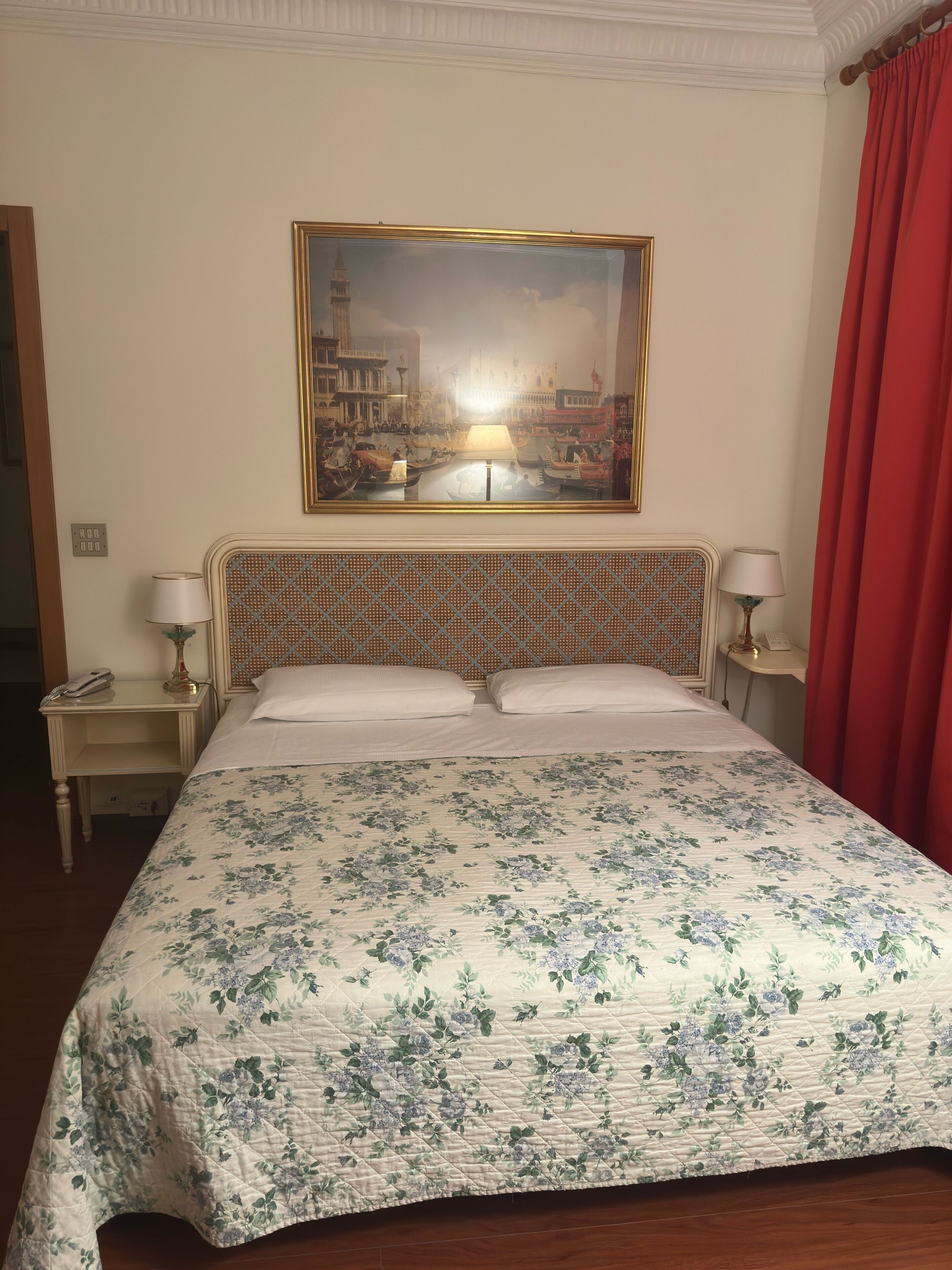 A hotel room with a bed, a painting of a cityscape, a nightstand with a lamp, and a red curtain.