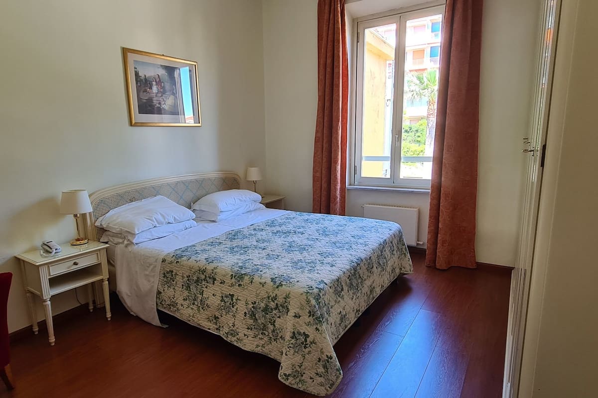 Een slaapkamer met een bed, een nachtkastje, een stoel, een raam met gordijnen en een schilderij aan de muur.