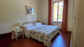 Un dormitorio con cama, mesita de noche, silla, ventana con cortinas y un cuadro en la pared.