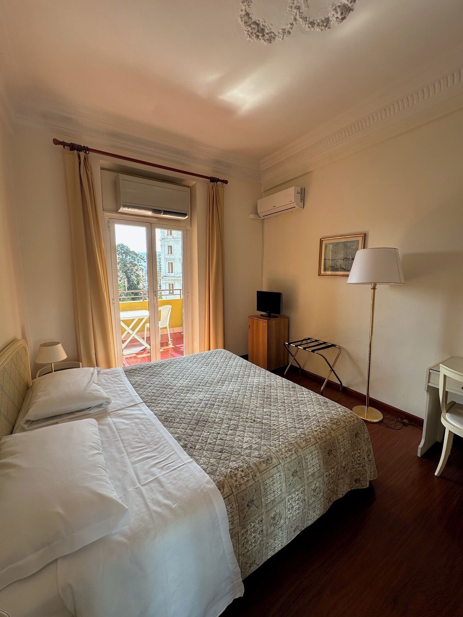 A hotel room with a bed, a desk, an air conditioning unit, a window with curtains, and a view of the outside.
