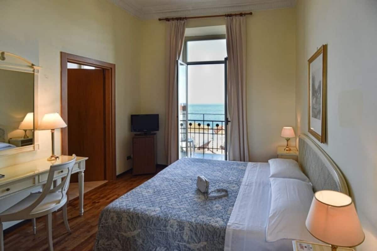 Een hotelkamer met een bed, een bureau, een stoel, een televisie en een balkon met uitzicht op zee.