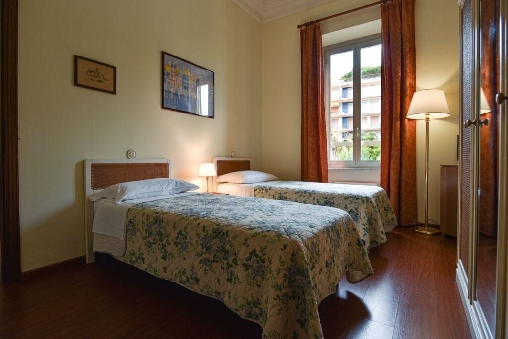 A hotel room with two beds, a window with curtains, a lamp, and framed pictures on the wall.