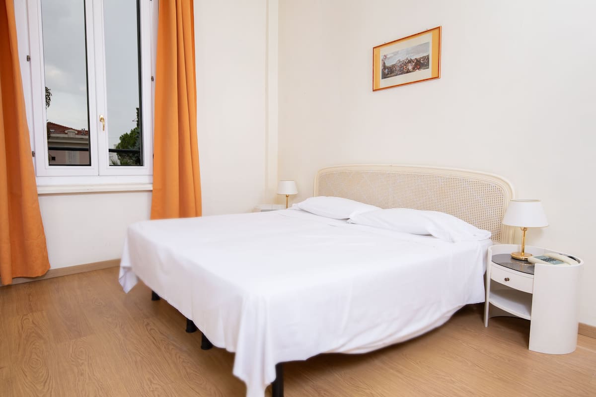 Een netjes opgemaakt bed met witte lakens, een nachtkastje met een lamp en een raam met gordijnen.