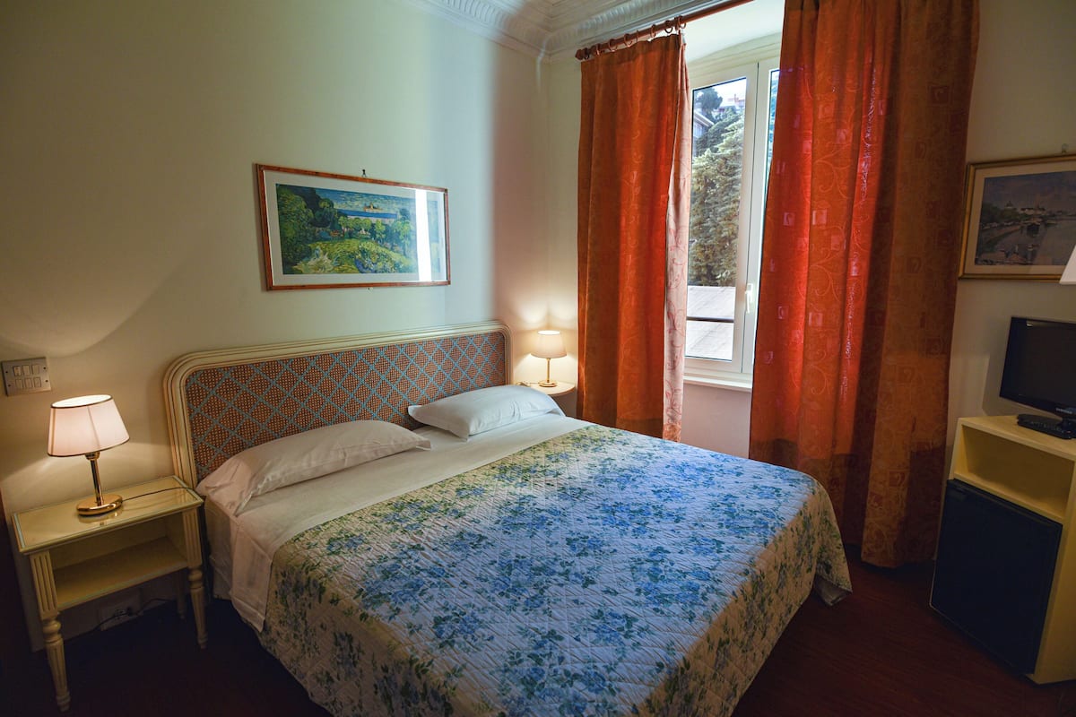 Een slaapkamer met een bed, nachtkastje met lamp, televisie op een dressoir en een raam met gordijnen.