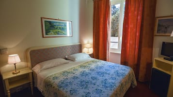 Un dormitorio con una cama, una mesita de noche con lámpara, un televisor sobre un mueble y una ventana con cortinas.