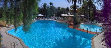 Una piscina con agua cristalina azul, rodeada de sillas de descanso y sombrillas, con palmeras al fondo.