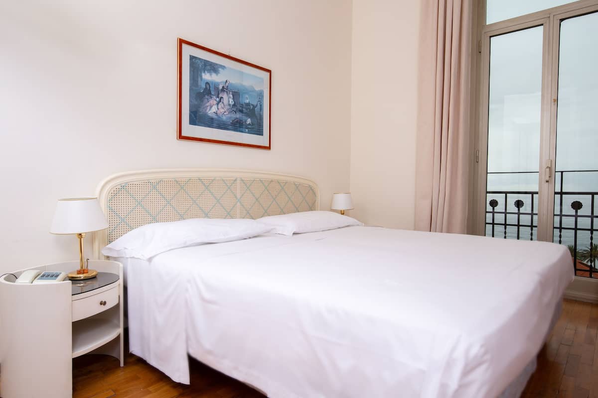 Een netjes opgemaakt bed met witte lakens, een nachtkastje met een lamp, een ingelijste foto aan de muur en een balkon met een hekwerk.