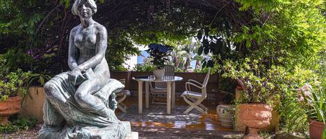 Une statue représentant une femme assise sur un piédestal, dans un jardin agrémenté d’une table et de chaises, entourée de plantes en pot et d’une pergola.