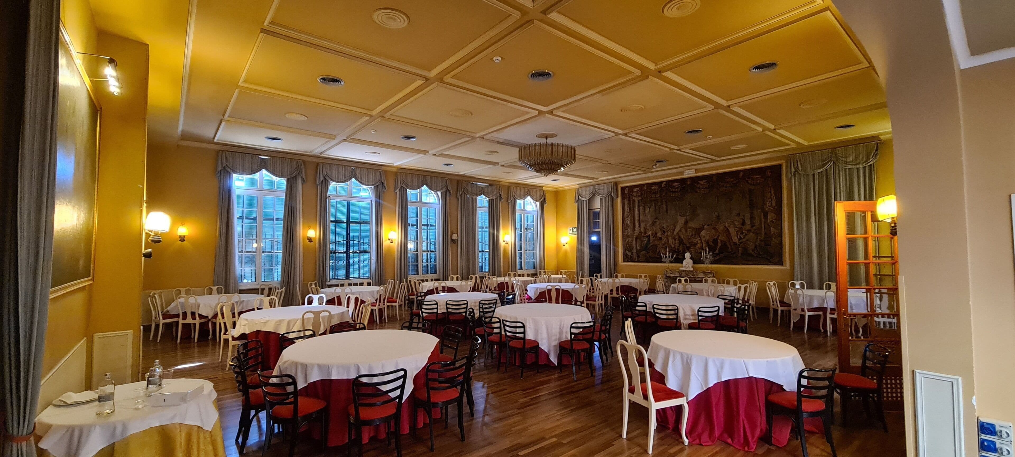 A dining area with round tables and chairs, large windows, and a chandelier.