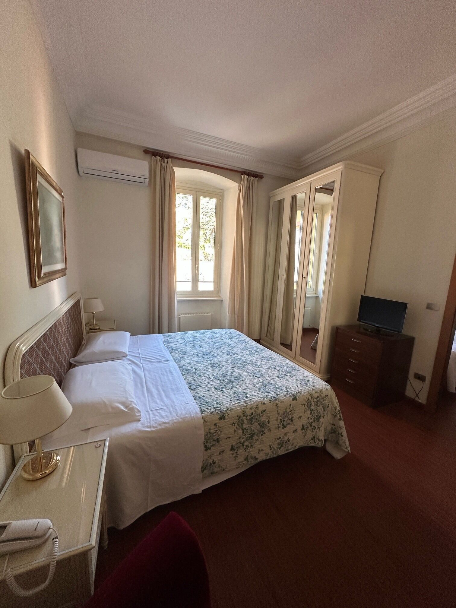 A hotel room with a bed, a desk with a lamp, a TV on a dresser, and a window with curtains.