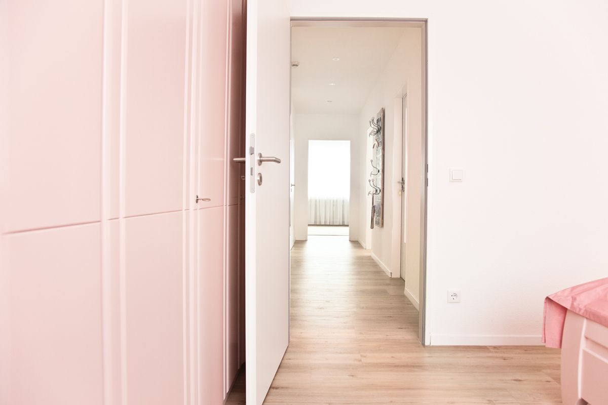 Un pasillo con suelo de madera, una puerta a la izquierda y una cama con cubierta rosa a la derecha.