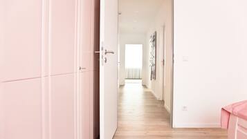 Un pasillo con suelo de madera, una puerta a la izquierda y una cama con cubierta rosa a la derecha.