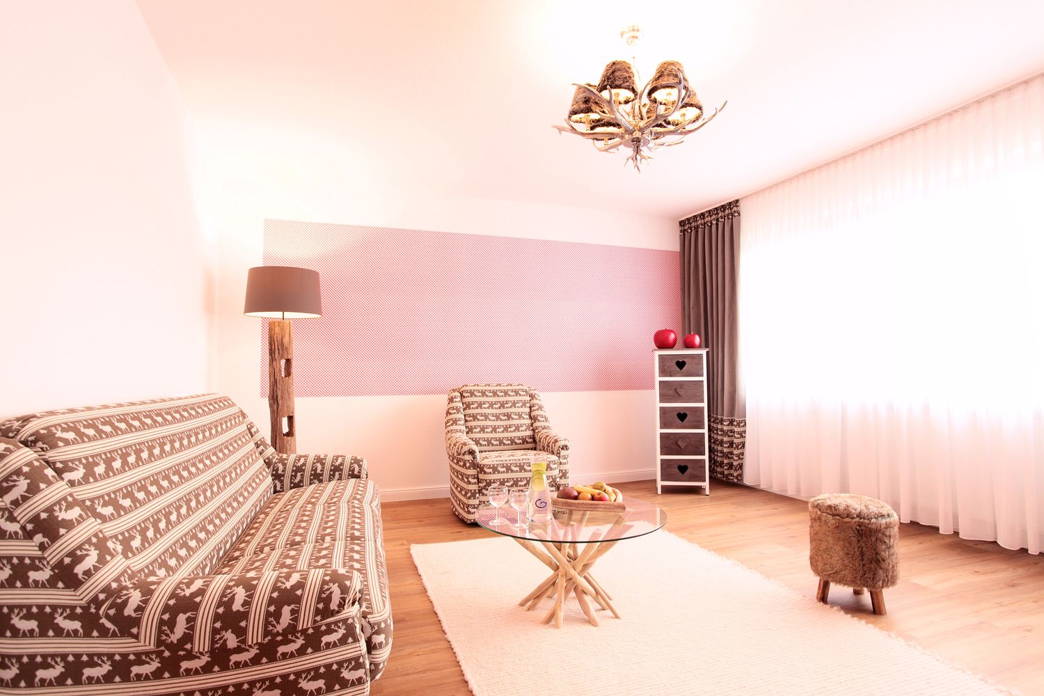 A living room with a patterned sofa, a small round table with a glass top, a patterned armchair, a wooden stool, a lamp, a ladder with storage drawers, and a chandelier.