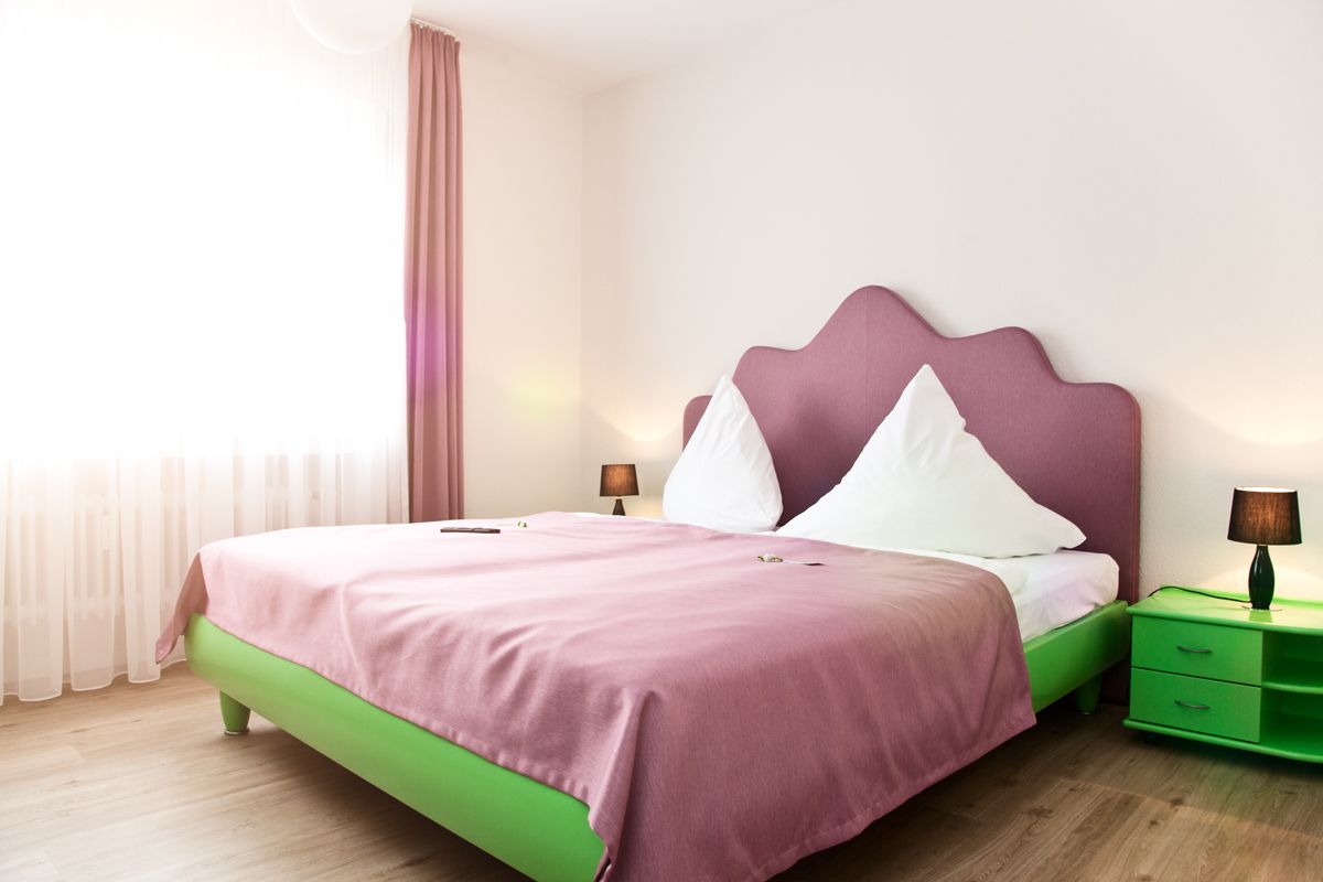 A neatly made bed with a pink and green color scheme, a bedside table with a lamp, and a window with sheer curtains.