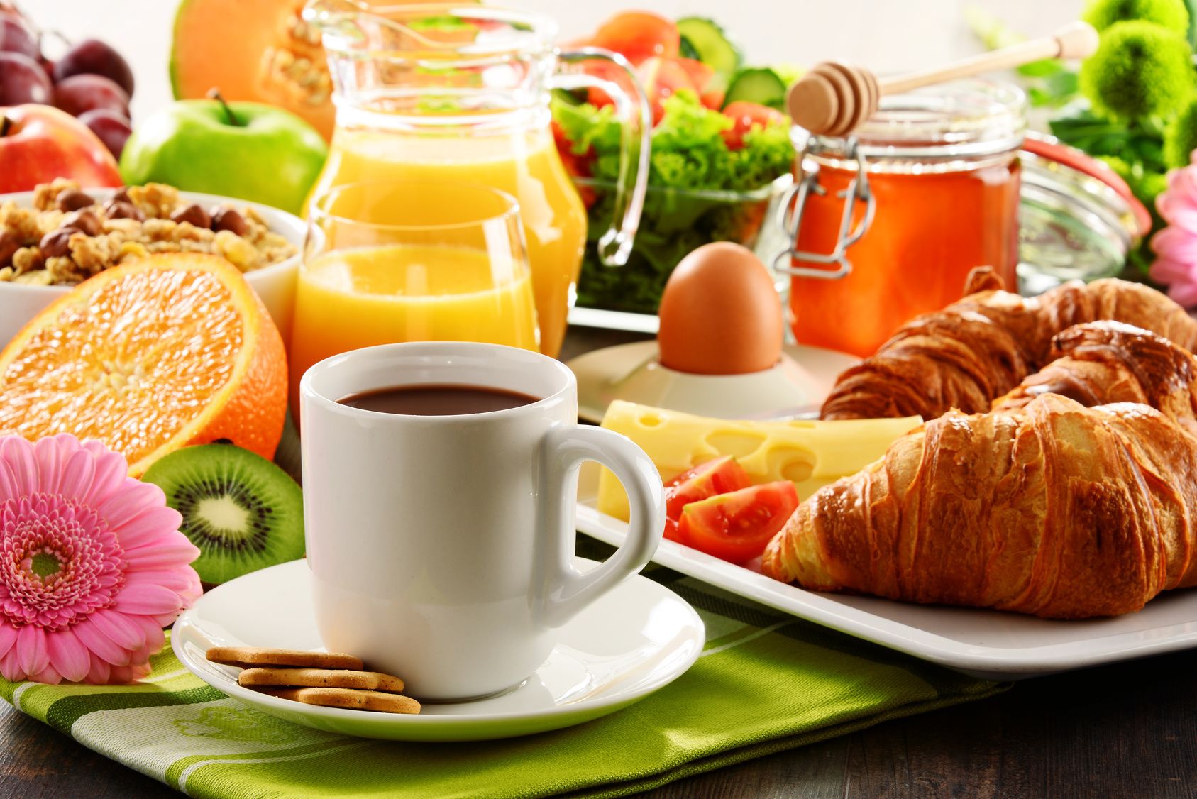 Ein Frühstückstisch mit einer Kaffeetasse, Croissants, Käse, Obst und einem Honigglas.