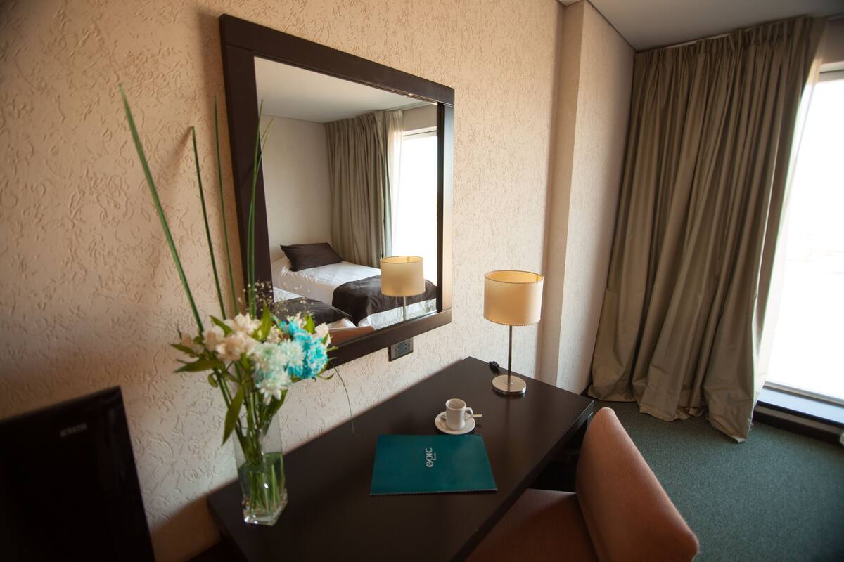 Ein Hotelzimmer mit einem Bett, einem Schreibtisch mit Lampe, einer Vase mit Blumen und einem Fenster mit Vorhängen.