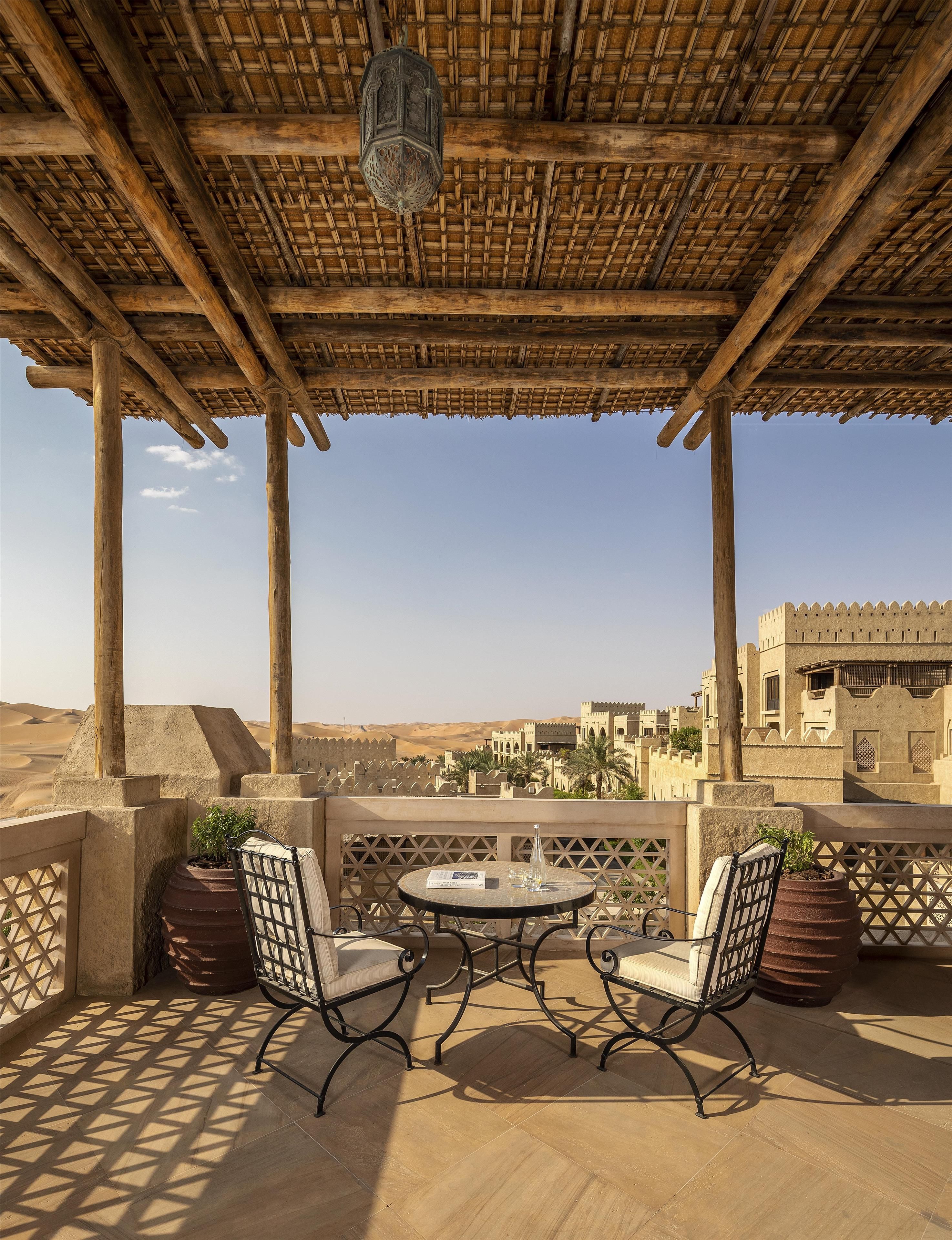 A terrace with a table and chairs, overlooking a desert landscape with traditional architecture.
