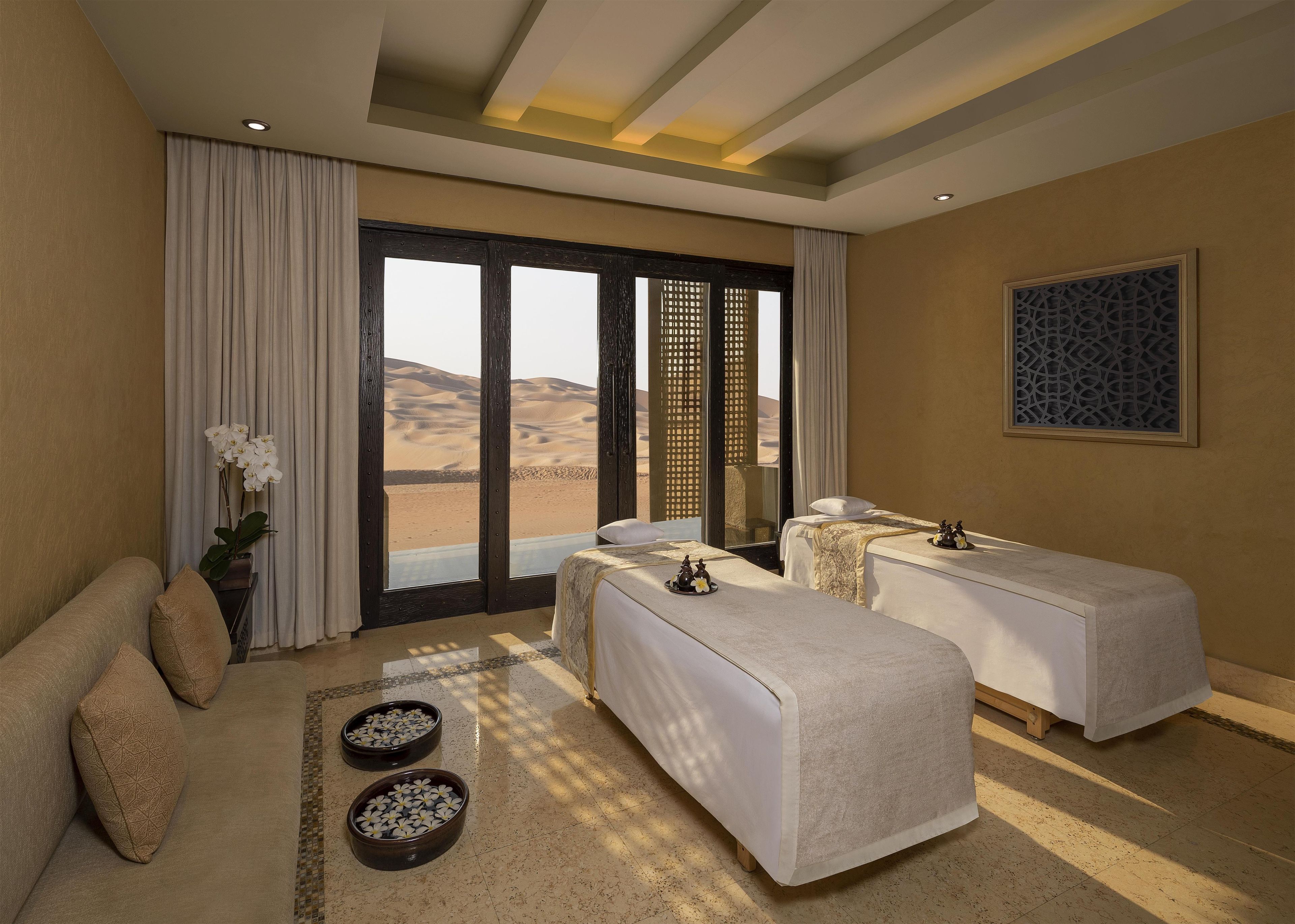 A hotel room with two beds, a sofa, a large window with a view of sand dunes, and a decorative wall piece.