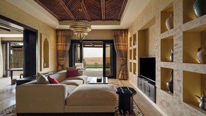 A spacious living room with a sofa, a coffee table, and a TV. There is a large window with curtains, a chandelier, and a patterned rug. - Qasr Al Sarab Desert Resort by Anantara (Al Mirayr)