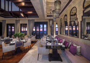 Un amplio comedor con varias mesas dispuestas para comer, candelabros elegantes y grandes ventanales con vistas al atardecer.
