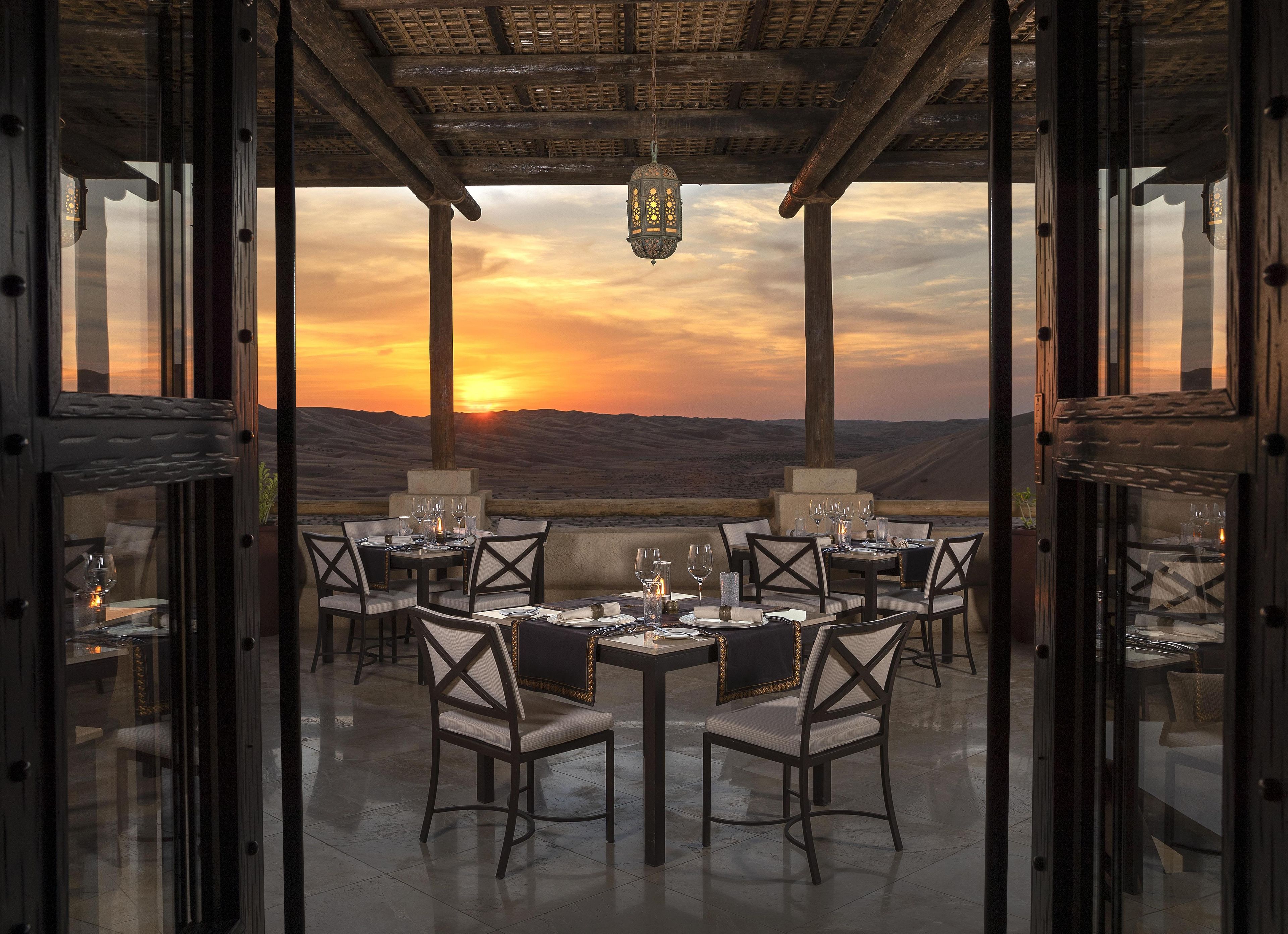 An outdoor dining area with tables and chairs, overlooking a desert landscape at sunset.