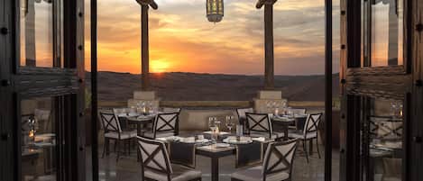 An outdoor dining area with tables and chairs, overlooking a desert landscape at sunset.