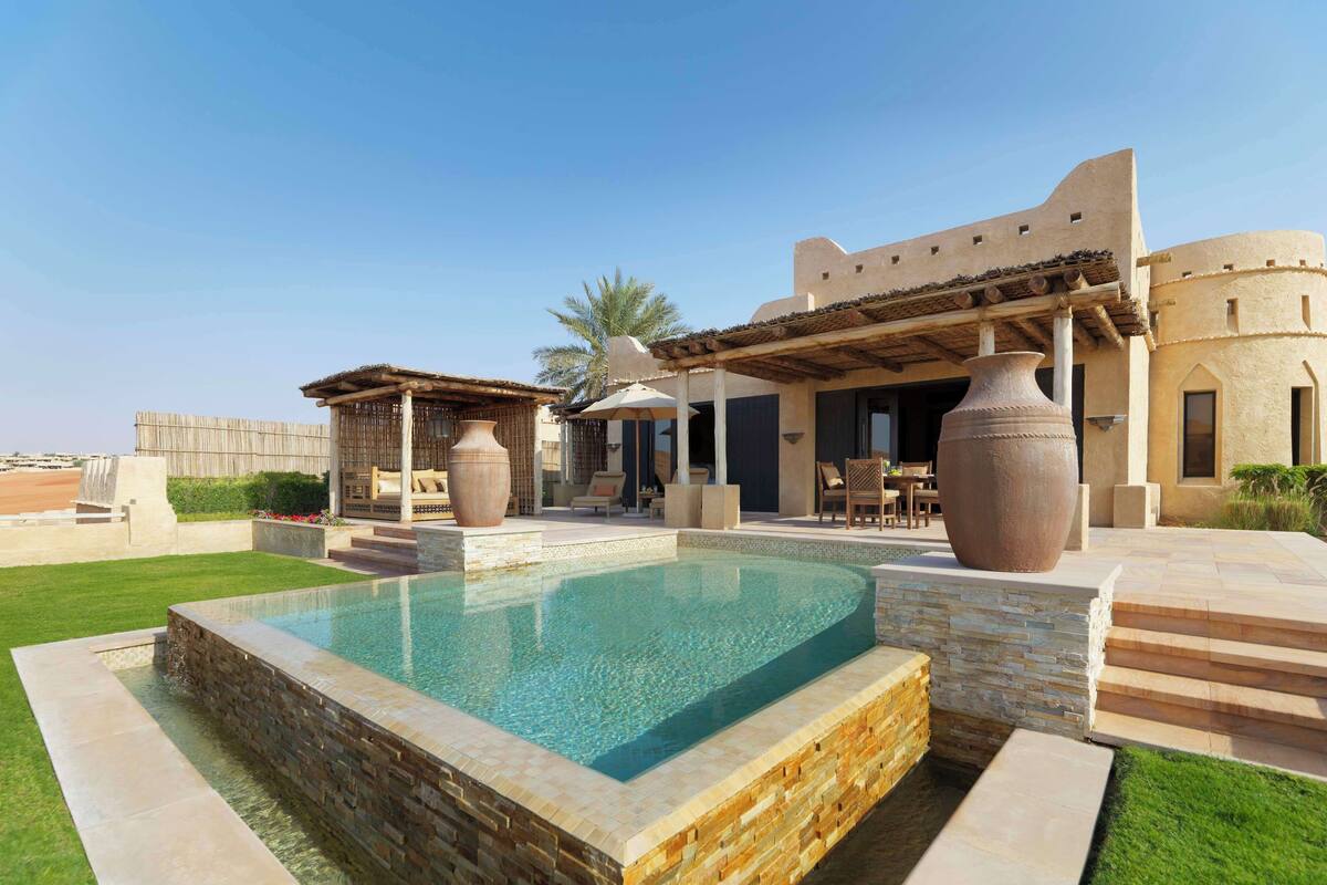 A pool area with a stone-lined pool, outdoor seating, and a building with a flat roof and arched windows.