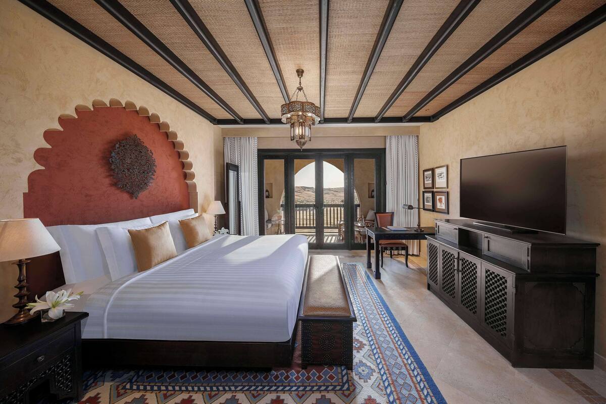 A hotel room with a large bed, a TV, a desk, and a view of the desert through the door.