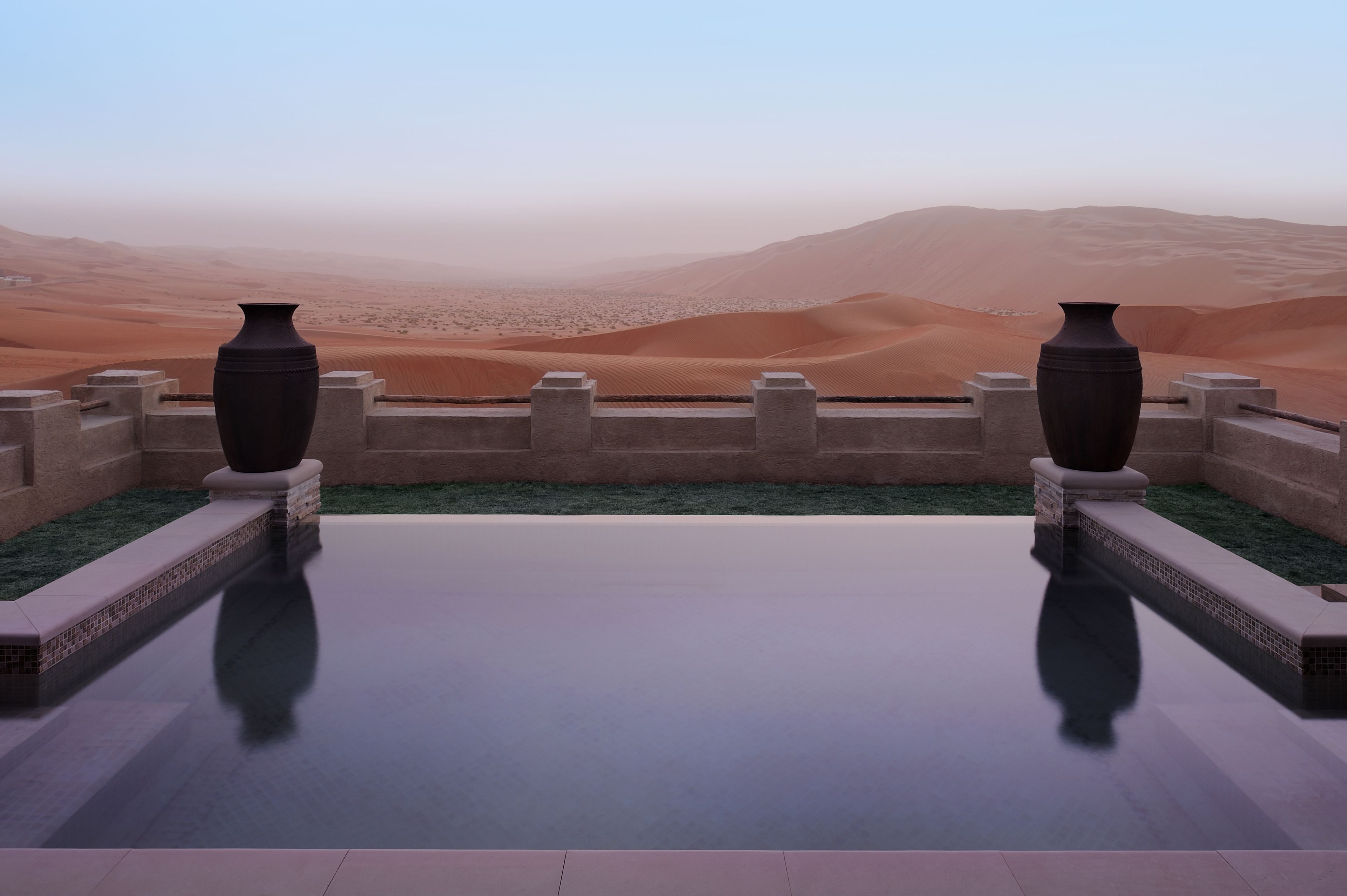 Ein Pool mit zwei großen Vasen an jeder Seite, umgeben von einer niedrigen Mauer und Blick auf Sanddünen.