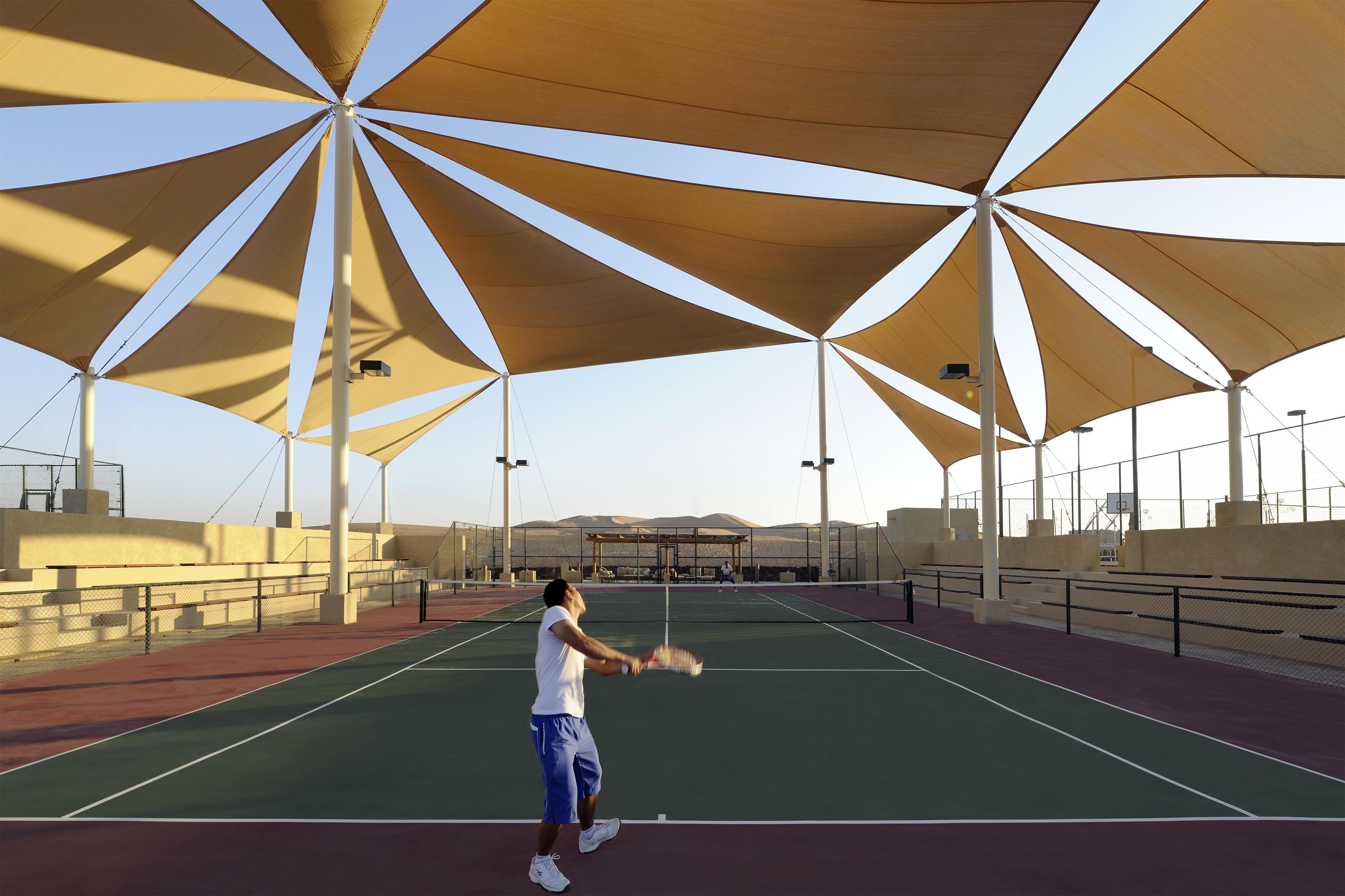 An outdoor tennis court with a net, surrounded by seating areas and shaded by large, triangular canopies.