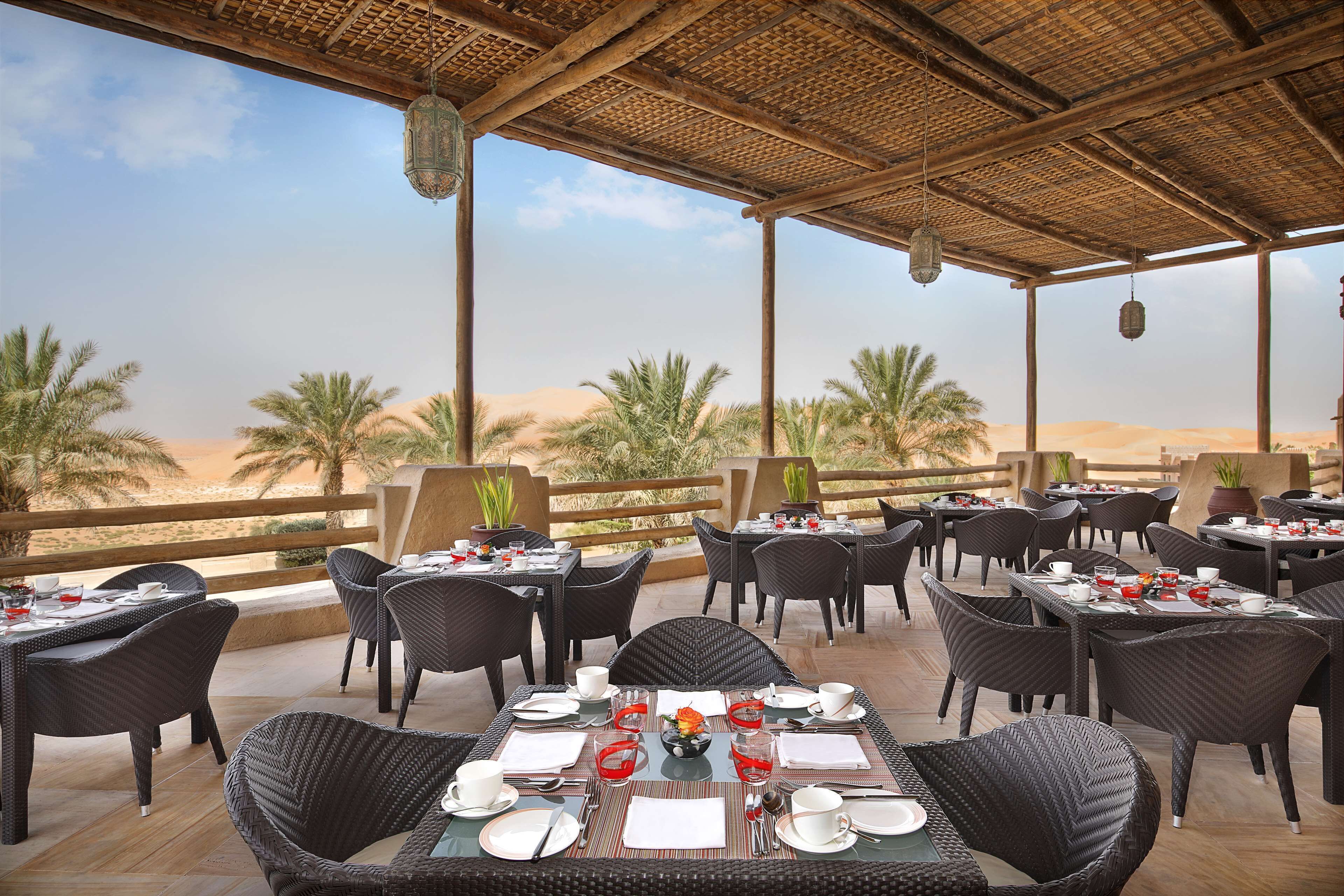An outdoor dining area with tables and chairs, overlooking a desert landscape with palm trees.