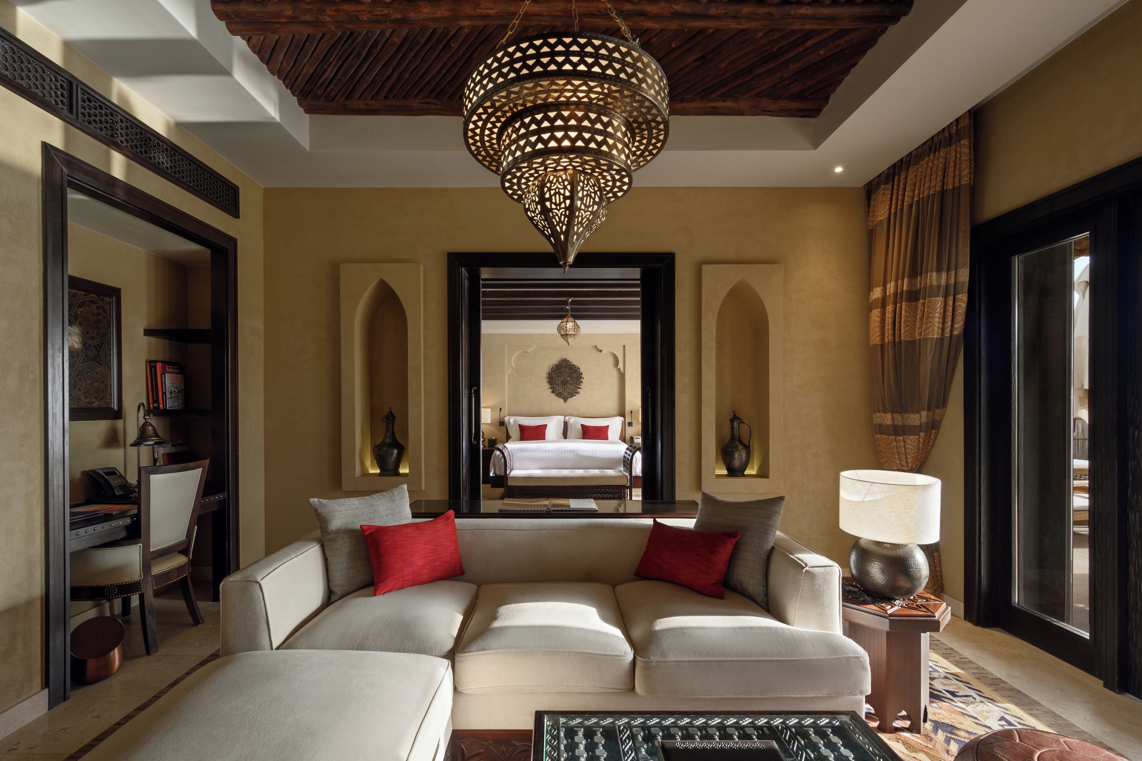 A modern living room with a large sectional sofa, a chandelier, and a view of a bedroom through an arched doorway.