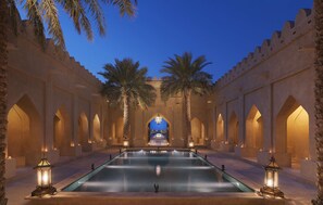 Un área de piscina con una fuente, palmeras y paseos con arcos.