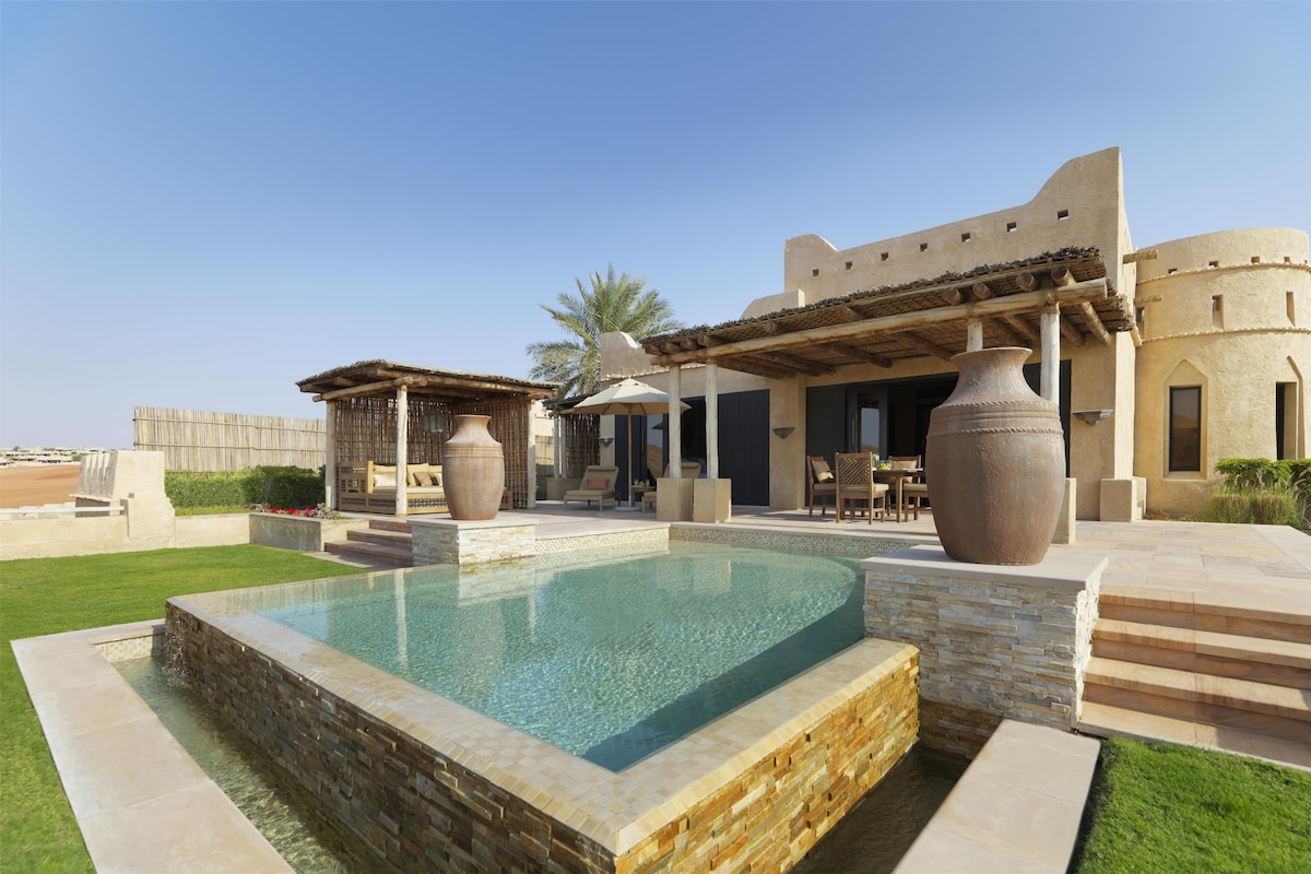 A pool area with a stone border, outdoor furniture, and a building with a flat roof.