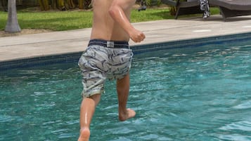 Zona de piscina con un niño jugando en el agua, rodeada de palmeras y sillas de descanso.