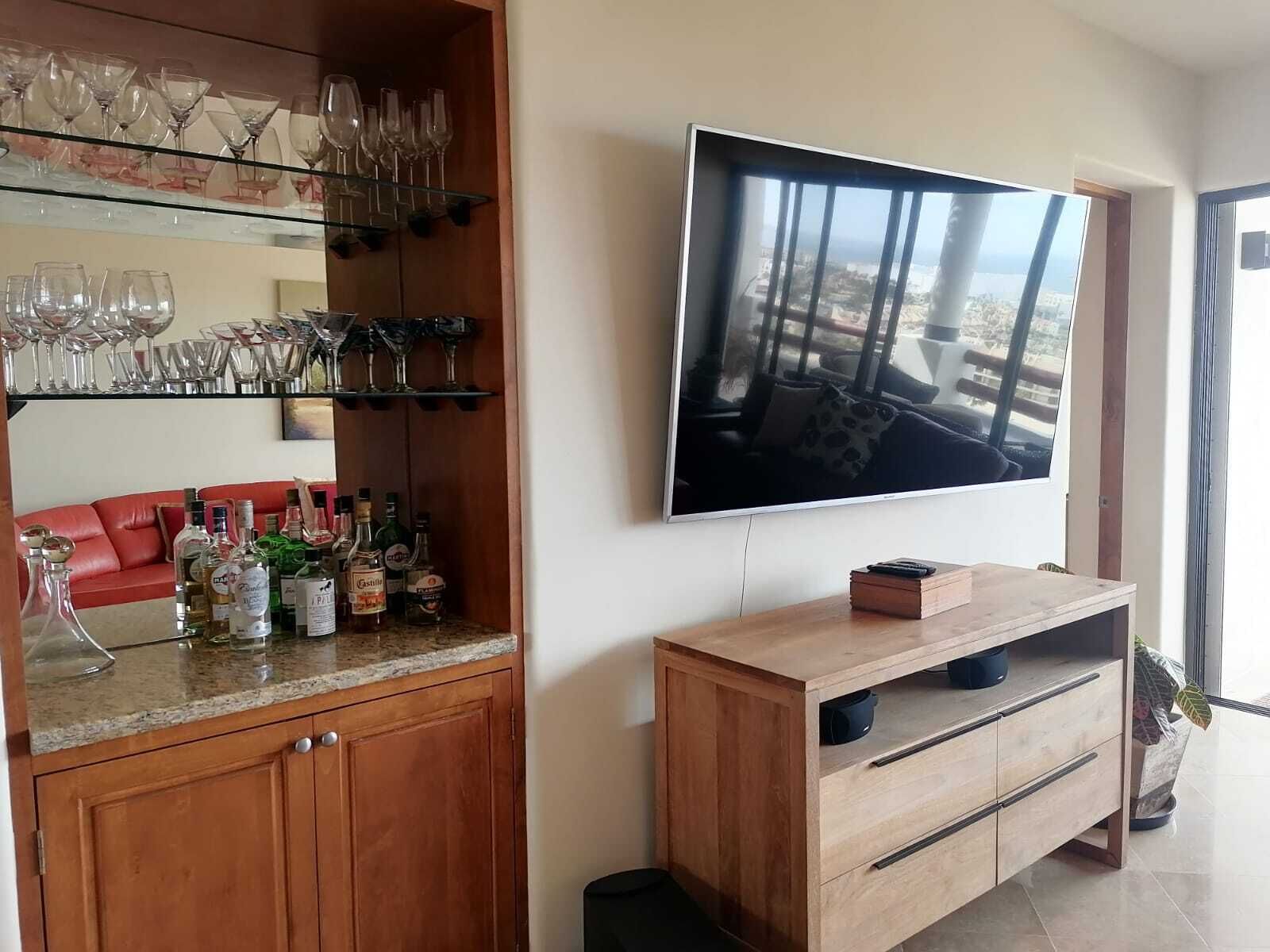A living room with a TV mounted on the wall, a wooden cabinet with bottles, and a red sofa.